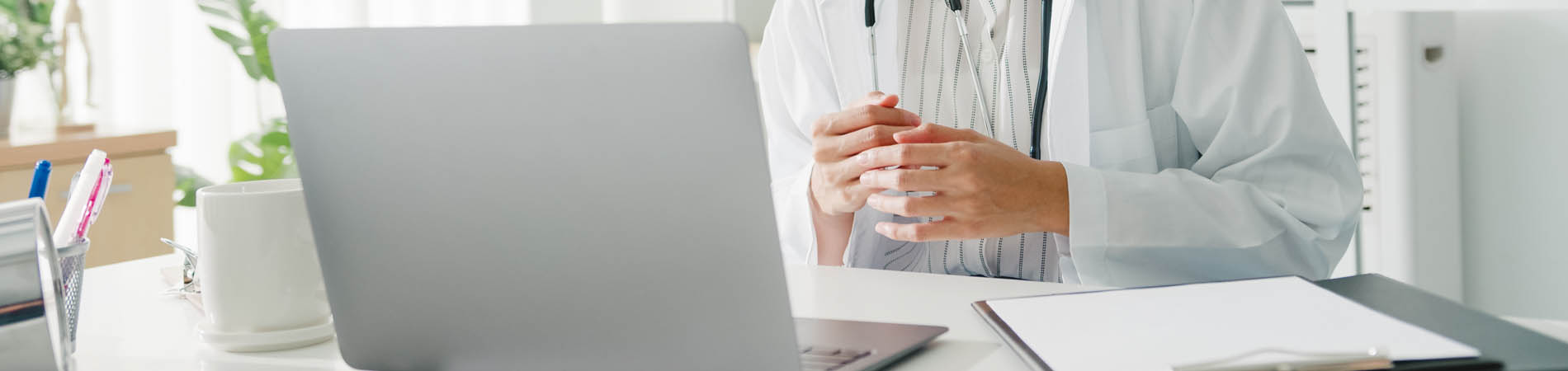 A doctor providing virtual care via a laptop in a white room with a notebook.